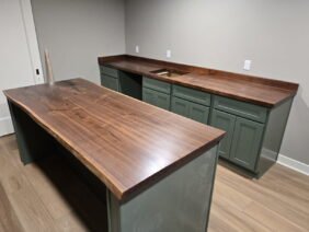 A kitchen with green cabinets and smooth, dark wooden countertops. An island with matching wood top and green base sits in the center. The room has light-colored walls and hardwood flooring.