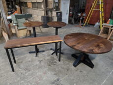 Four wooden tables with black metal legs—a round coffee table, a rectangular bench, and two small round side tables—are arranged in a workshop with tools, ladders, and industrial equipment in the background.