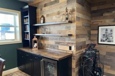 Home office with rustic wooden walls and shelves displaying books and decor. A wooden countertop has a mini-fridge below. The framed picture on the wall and a window with blinds complete the cozy setting.